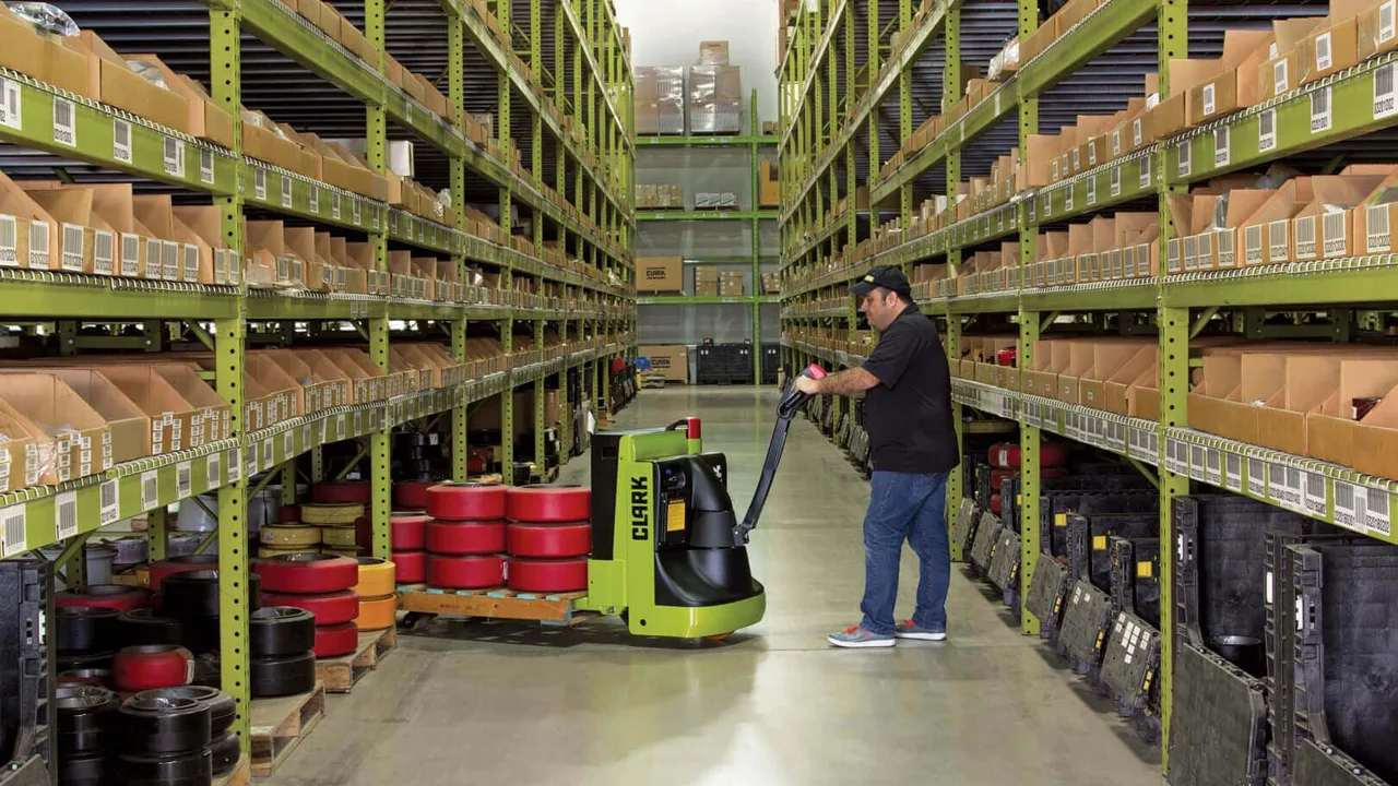 A lime green Clark electric pallet truck being driven down a stocked warehouse storage aisle, with Clark branding on the side